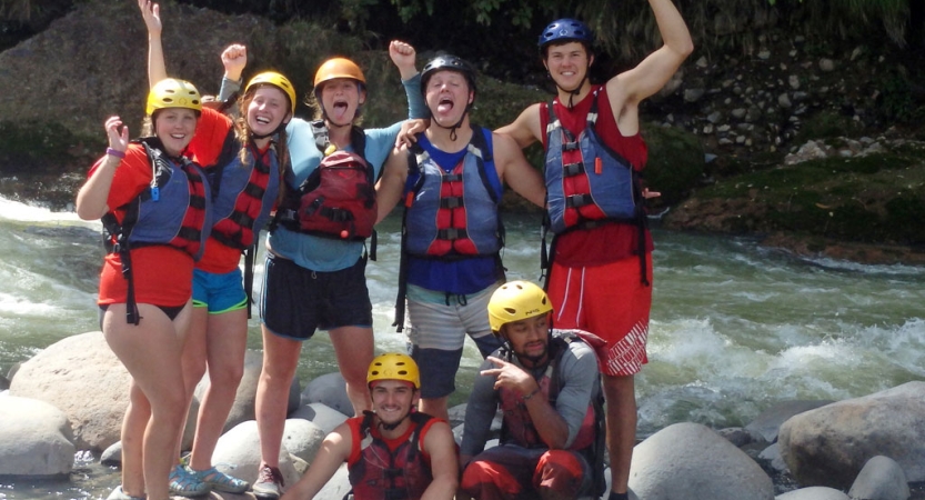 a group of people wearing safety gear stand on rocks among whitewater and pose for a group photo